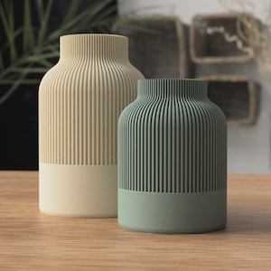Può includere: Due vasi decorativi con un design a coste. Uno è beige chiaro, l'altro verde tenue. Entrambi i vasi hanno una base in tinta unita. I vasi sono esposti su una superficie di legno.