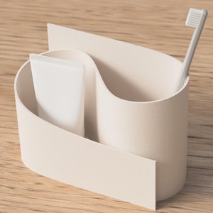 Può includere: Un portaspazzolino color crema con un design unico e fluido. Contiene un tubetto di dentifricio bianco e uno spazzolino bianco. Il supporto è posizionato su una superficie di legno chiaro, offrendo un'estetica minimalista per l'organizzazione del bagno.