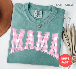 May include: Light green t-shirt with the word "MAMA" in pink and white plaid lettering. The shirt has a crew neck and short sleeves. A gold necklace with a pendant is draped across the shirt. The shirt is folded on a surface.