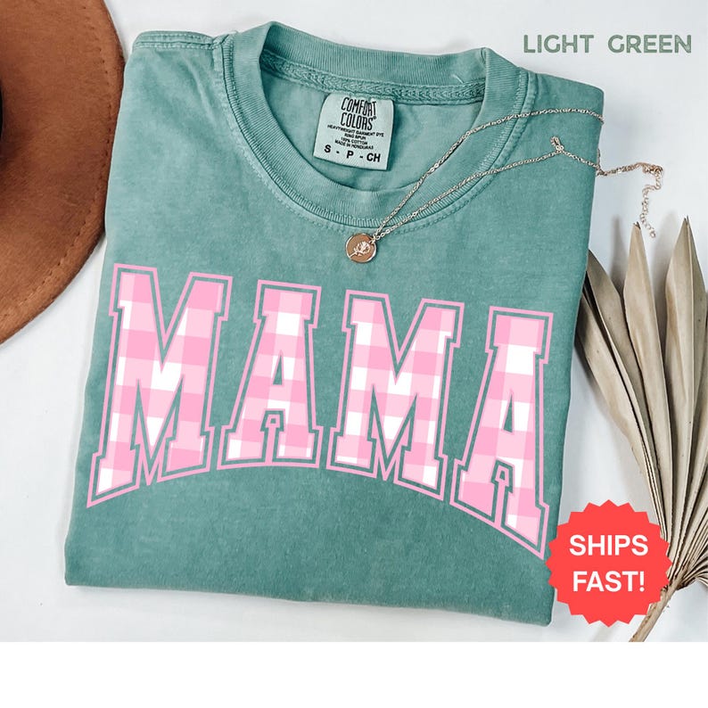 May include: Light green t-shirt with the word "MAMA" in pink and white plaid lettering. The shirt has a crew neck and short sleeves. A gold necklace with a pendant is draped across the shirt. The shirt is folded on a surface.