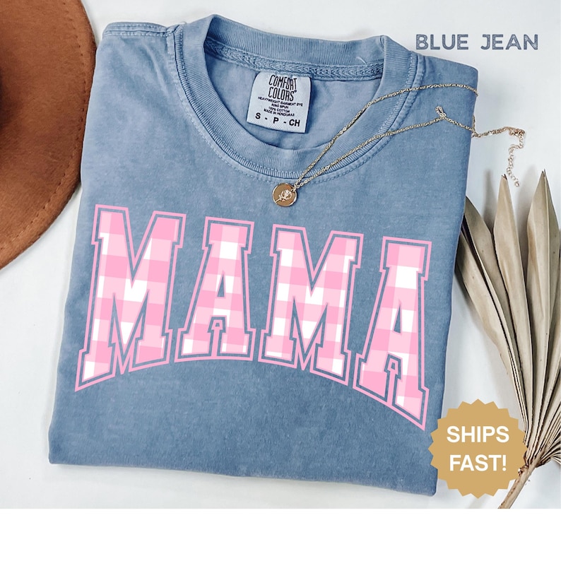 May include: Blue jean-coloured t-shirt with the word "MAMA" in large pink and white plaid letters. A gold necklace with a pendant is draped across the shirt. The shirt has a crew neck and short sleeves.