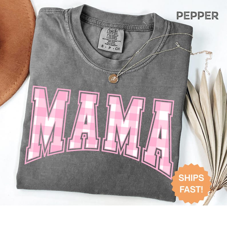 May include: A grey Comfort Colors t-shirt featuring the word "MAMA" in a pink and white plaid pattern. The shirt has a crew neck and short sleeves. A gold necklace with a pendant is draped across the shirt. The word "PEPPER" is printed at the top.