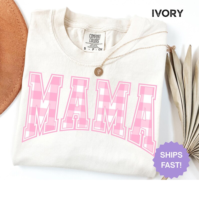 May include: Ivory-coloured t-shirt featuring the word "MAMA" in large, pink, gingham-patterned letters. The shirt has a crew neck and short sleeves. A gold necklace with a pendant is draped on the shirt. The shirt is labelled "Comfort Colors."