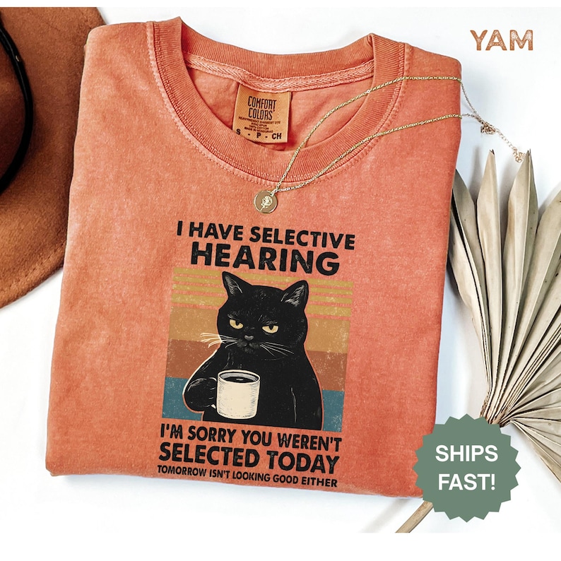 May include: Coral-colored Comfort Colors t-shirt with a graphic of a black cat holding a mug, and the text "I HAVE SELECTIVE HEARING" and "I'M SORRY YOU WEREN'T SELECTED TODAY". A gold necklace and a decorative leaf are also visible.