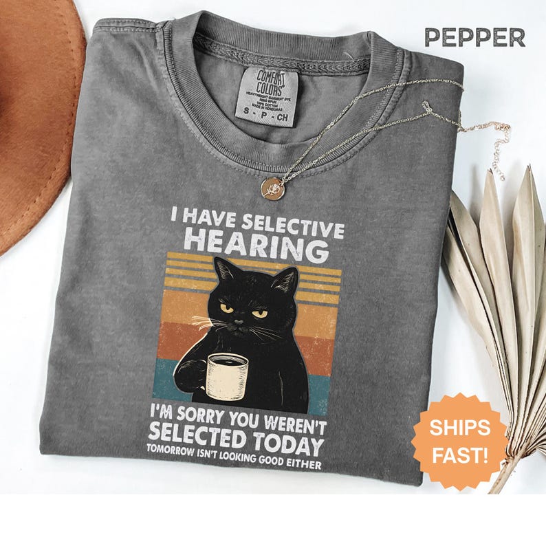 May include: Pepper-colored t-shirt with the text "I HAVE SELECTIVE HEARING" and a graphic of a black cat holding a mug. The shirt also says "I'M SORRY YOU WEREN'T SELECTED TODAY" and "TOMORROW ISN'T LOOKING GOOD EITHER". A gold necklace is draped on the shirt.