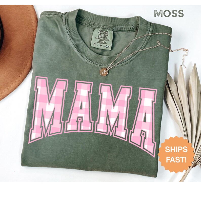 May include: A moss green t-shirt featuring the word "MAMA" in pink and white checked lettering. A gold necklace with a pendant is placed on the shirt. The t-shirt is folded on a white surface.