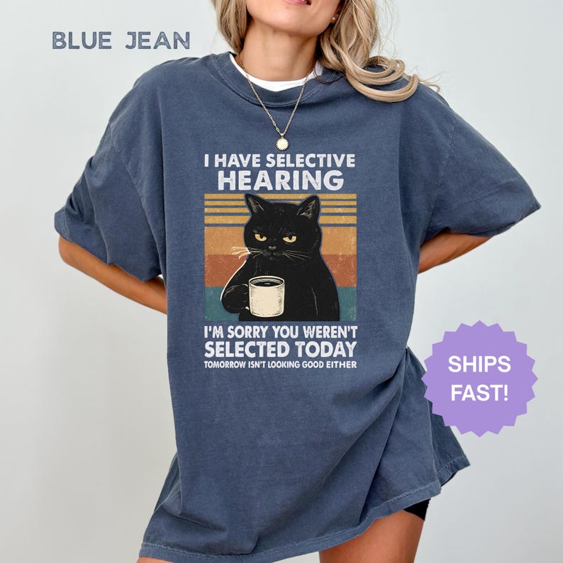 May include: Blue jean colored t-shirt with a graphic of a black cat holding a coffee cup. The text reads "I HAVE SELECTIVE HEARING" and "I'M SORRY YOU WEREN'T SELECTED TODAY". The shirt also has the text "TOMORROW ISN'T LOOKING GOOD EITHER".