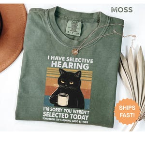 May include: Moss green t-shirt with the text "I HAVE SELECTIVE HEARING" and a graphic of a black cat holding a mug. The shirt also says "I'M SORRY YOU WEREN'T SELECTED TODAY". A gold necklace is draped across the shirt.