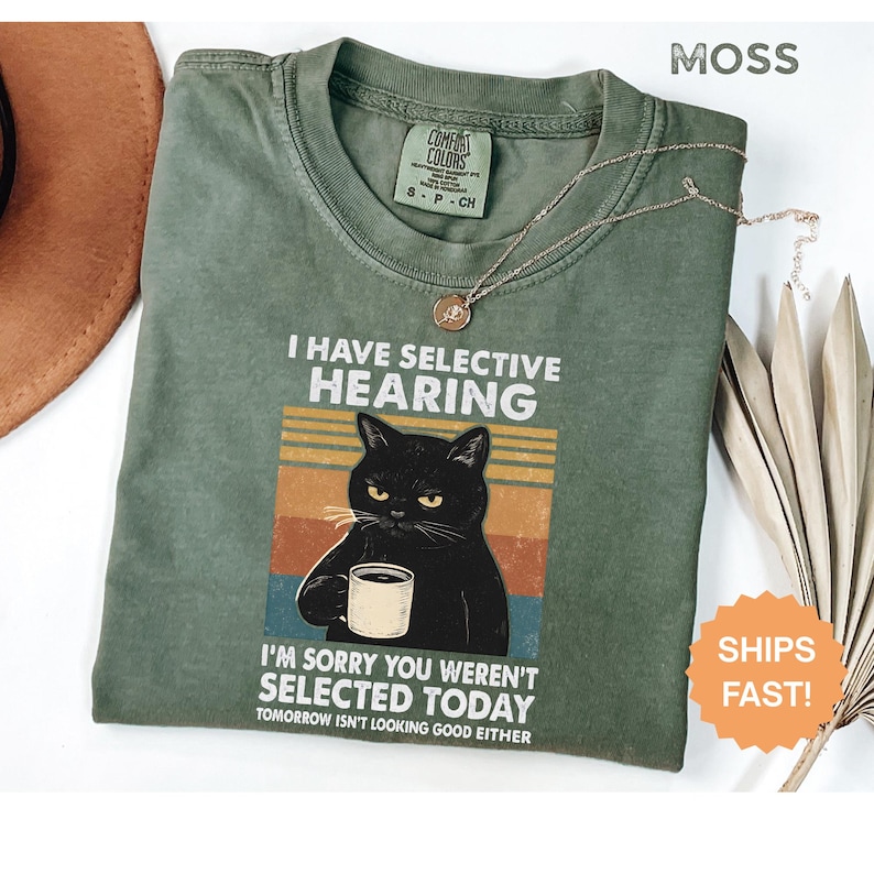 May include: Moss green t-shirt with the text "I HAVE SELECTIVE HEARING" and a graphic of a black cat holding a mug. The shirt also says "I'M SORRY YOU WEREN'T SELECTED TODAY". A gold necklace is draped across the shirt.