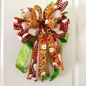 May include: A decorative bow featuring multiple ribbon patterns and colors. The bow includes ribbons with pumpkin and floral patterns, red and white gingham, and solid green glitter ribbon. The bow is likely for seasonal or holiday decor.