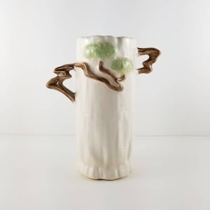 May include: White ceramic vase with two green leaves and brown branches forming handles. The vase has a textured surface and a scalloped rim.