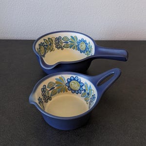 May include: Two navy blue ceramic sauce bowls with handles. The bowls have a cream-colored interior and are decorated with a floral pattern in shades of blue, green, and yellow. The bowls are designed for serving sauces or dips.