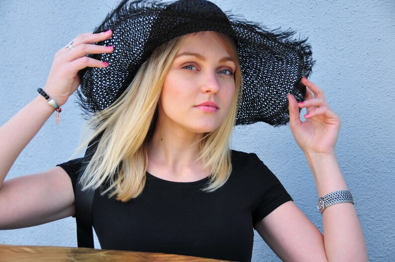 K&ouml;nnte beinhalten: Eine Frau mit blonden Haaren tr&auml;gt einen schwarzen breitkrempigen Sonnenhut mit einem schwarz-wei&szlig;en Muster. Sie tr&auml;gt ein schwarzes, kurz&auml;rmeliges Hemd.