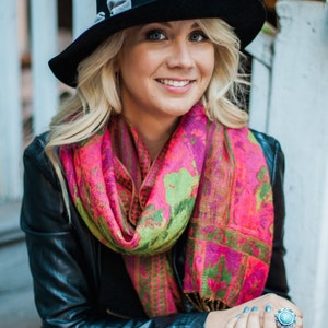 Peut inclure: Une femme portant un fedora noir avec un ruban gris, une &eacute;charpe &agrave; motifs roses et verts, une veste en cuir noir et un pantalon blanc. Elle est assise sur un escalier et porte une bague turquoise &agrave; son doigt.