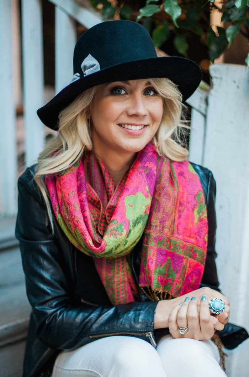 Peut inclure: Une femme portant un fedora noir avec un ruban gris, une &eacute;charpe &agrave; motifs roses et verts, une veste en cuir noir et un pantalon blanc. Elle est assise sur un escalier et porte une bague turquoise &agrave; son doigt.