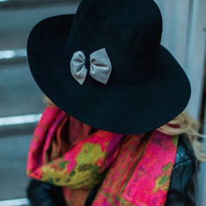 Peut inclure: Un chapeau fedora en feutre noir &agrave; large bord et un n&oelig;ud en velours gris. Le chapeau est port&eacute; par une personne portant une &eacute;charpe color&eacute;e et une veste en cuir noir.