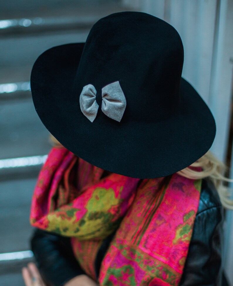 Peut inclure: Un chapeau fedora en feutre noir &agrave; large bord et un n&oelig;ud en velours gris. Le chapeau est port&eacute; par une personne portant une &eacute;charpe color&eacute;e et une veste en cuir noir.