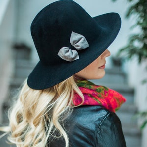 Peut inclure: Un chapeau fedora en feutre noir &agrave; large bord et un n&oelig;ud argent&eacute;. Le chapeau est port&eacute; par une personne aux longs cheveux blonds et une &eacute;charpe color&eacute;e.