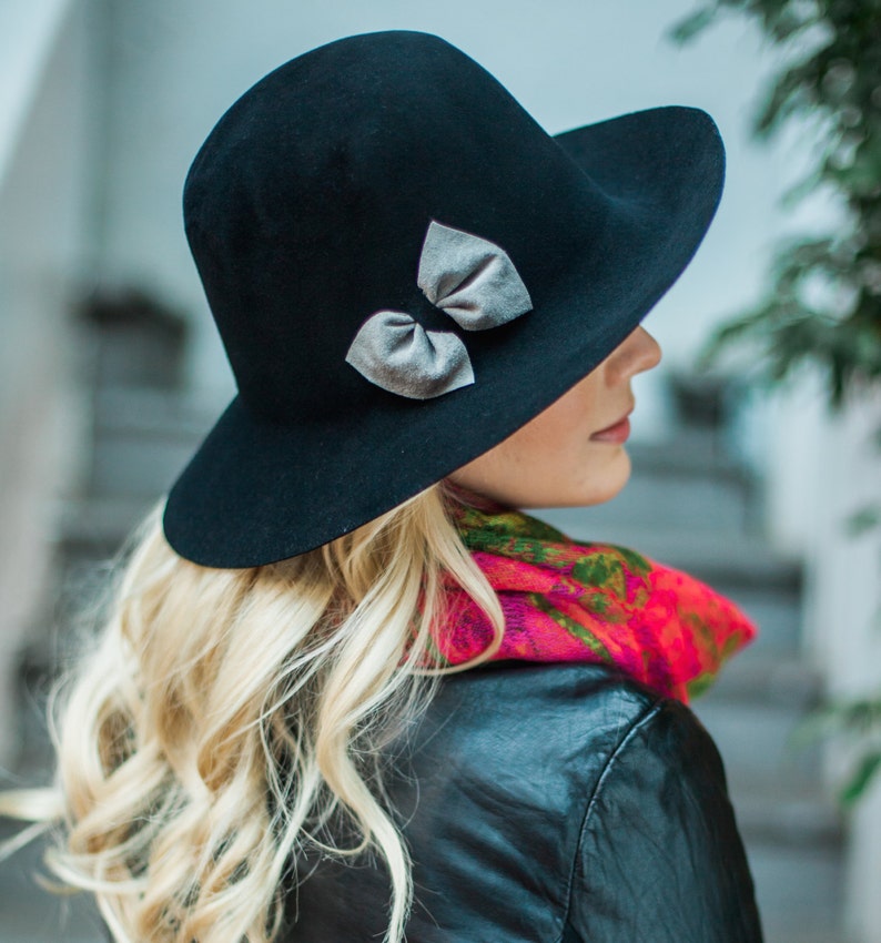 Peut inclure: Un chapeau fedora en feutre noir &agrave; large bord et un n&oelig;ud argent&eacute;. Le chapeau est port&eacute; par une personne aux longs cheveux blonds et une &eacute;charpe color&eacute;e.