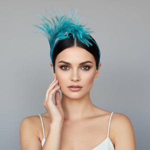 May include: A teal feather headband with a matching band. The headband features a cluster of upright feathers on the left side. The model is wearing a white spaghetti strap top.