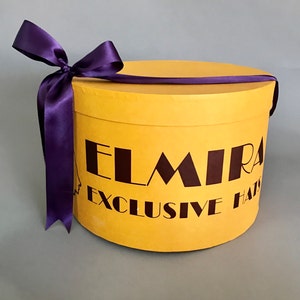 May include: A yellow hat box with a purple ribbon and the text "ELMIRA EXCLUSIVE HATS" printed on the side.