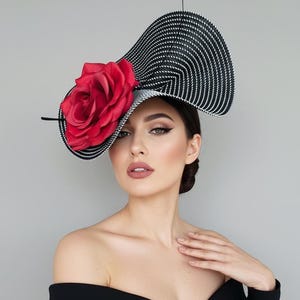 Könnte beinhalten: Ein Hut mit schwarz-weißem Streifenmuster und einer großen roten Rose. Der Hut hat eine einzigartige, asymmetrische Form und ist wahrscheinlich für einen besonderen Anlass gedacht. Ein auffälliges Modeaccessoire.