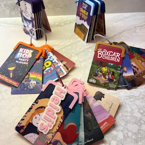 May include: A collection of colourful book tags, some with titles like "Charlie and the Chocolate Factory" and "The Boxcar Children." The tags have pink, orange, and blue accents. One tag features the name "Heidi's" in pink.