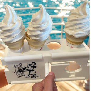May include: Three soft-serve ice cream cones in a white holder with a Mickey Mouse graphic. The holder has a design of Mickey Mouse steering a ship's wheel. The background is a blurred view of the ocean.