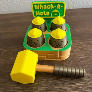 May include: A yellow and brown Whack-A-Mole game with a matching yellow mallet. The game features four brown moles wearing yellow hard hats. The sign on top reads "Whack-A-Mole".