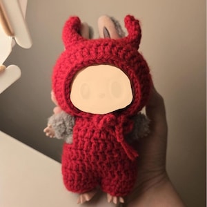 May include: A handmade red crochet doll wearing a devil-horned hood and matching jumpsuit. The doll has gray fur-like arms and a neutral face. The doll is held in a hand.