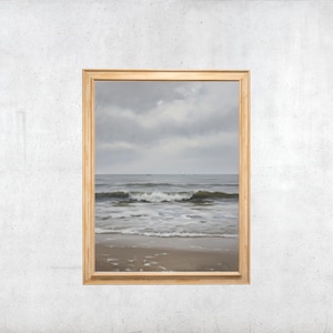 Peut inclure: Une impression d'art encadrée représentant un paysage marin avec une plage de sable, des vagues douces se brisant sur le rivage et un océan calme sous un ciel gris nuageux. L'œuvre est insérée dans un cadre en bois clair au profil simple.