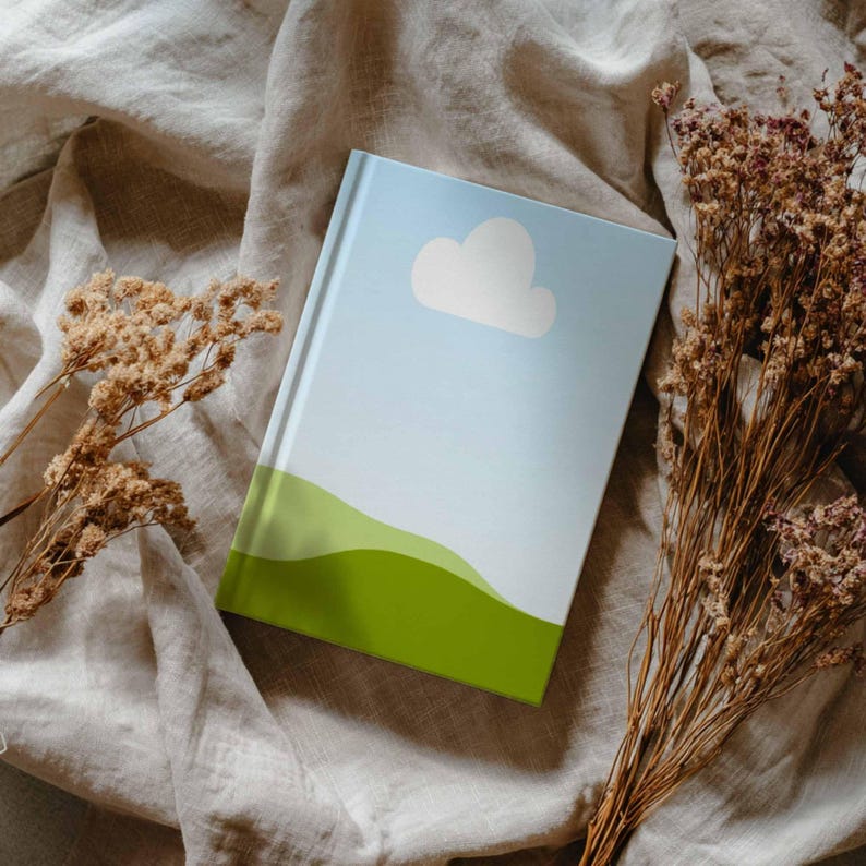 May include: A hardcover journal with a sky blue cover featuring a white cloud and green rolling hills. The journal is resting on a beige linen surface, with dried flowers on either side.