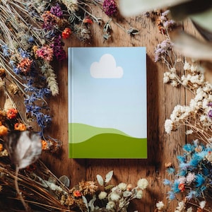 May include: A hardcover notebook with a sky and cloud design. The cover features a light blue sky, a white cloud, and green rolling hills. The notebook is surrounded by dried flowers on a wooden surface.