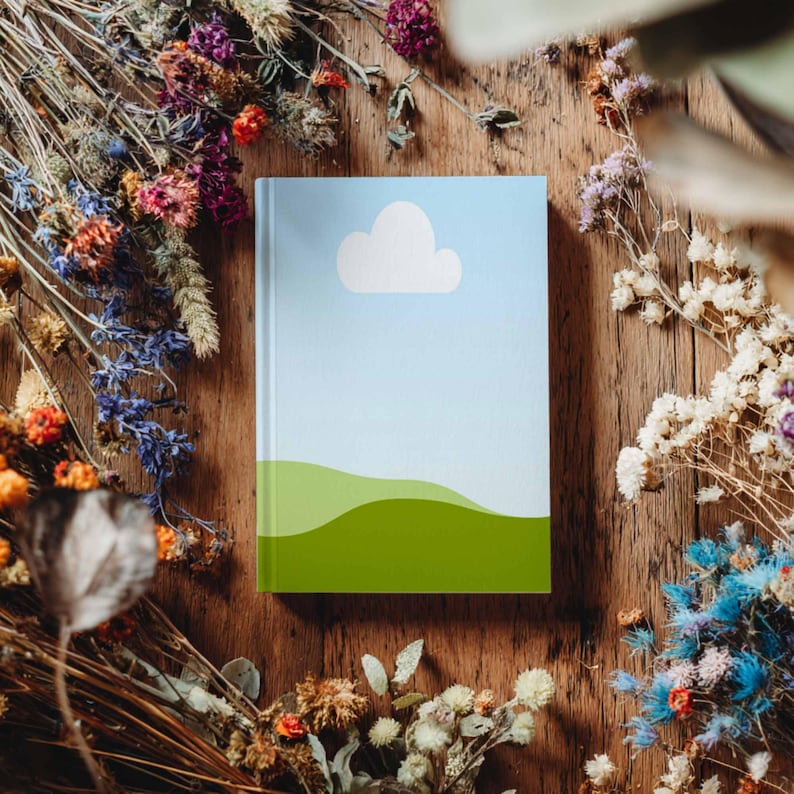 May include: A hardcover notebook with a sky and cloud design. The cover features a light blue sky, a white cloud, and green rolling hills. The notebook is surrounded by dried flowers on a wooden surface.