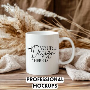 May include: A white ceramic mug with a curved handle, featuring the text "YOUR Design HERE" in a stylish script. The mug is set against a neutral background with dried plants and fabric, with the text "PROFESSIONAL MOCKUPS" at the bottom.