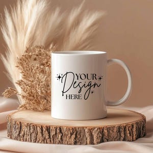 May include: A white ceramic mug with the text "YOUR Design HERE" in black script. The mug sits on a wooden slice, with dried ornamental grass in the background. The mug has a curved handle.