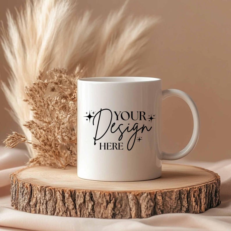 May include: A white ceramic mug with the text "YOUR Design HERE" in black script. The mug sits on a wooden slice, with dried ornamental grass in the background. The mug has a curved handle.