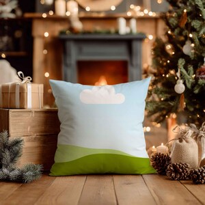 May include: Square cushion with a sky and landscape design. The cushion features a light blue background with a white cloud and green rolling hills at the bottom. The cushion is on a wooden surface.
