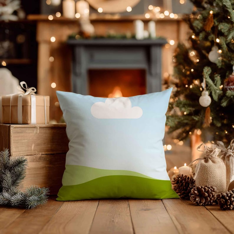 May include: Square cushion with a sky and landscape design. The cushion features a light blue background with a white cloud and green rolling hills at the bottom. The cushion is on a wooden surface.