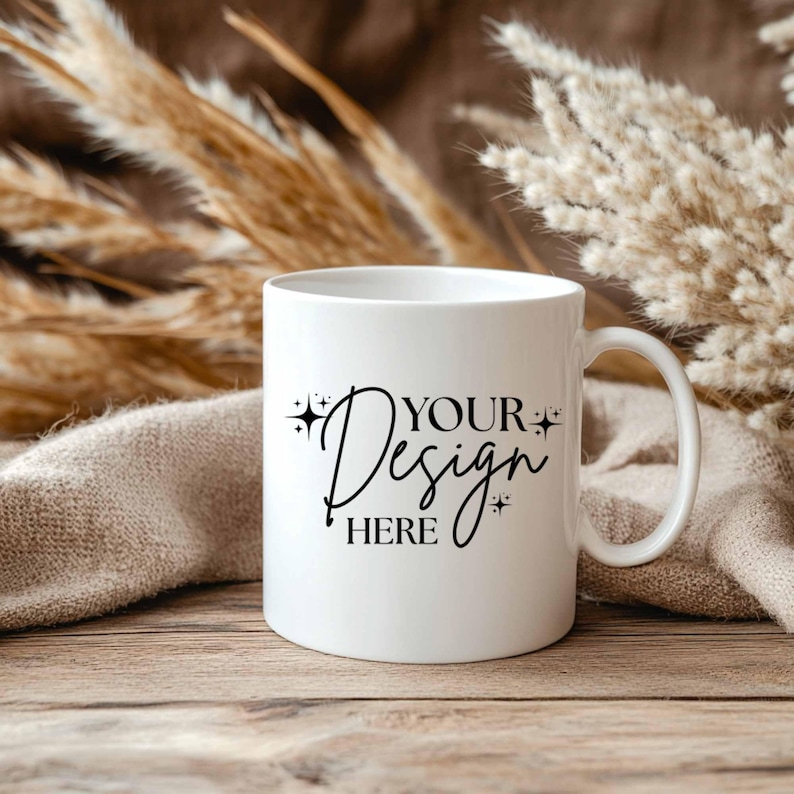 May include: A white ceramic mug with a curved handle. The mug features the text "YOUR Design HERE" in a black script font. The mug is set on a wooden surface with dried plants and a beige cloth in the background.