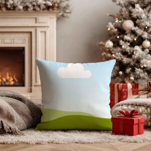 May include: Square cushion featuring a light blue sky design, a white cloud, and green rolling hills. The cushion is placed on a light-coloured rug, with a Christmas tree and fireplace in the background.