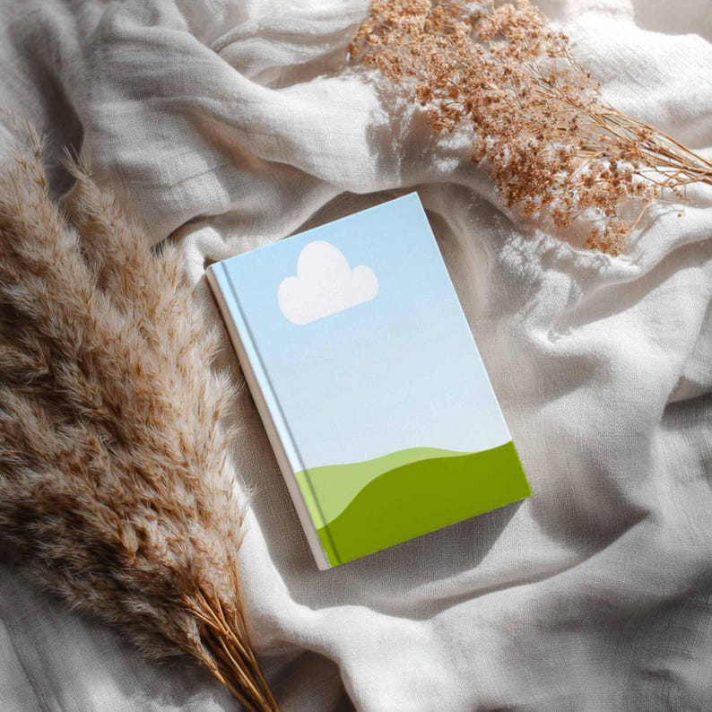 May include: A closed notebook with a blue sky and cloud design on the cover. The bottom of the cover has a green hill design. The notebook is lying on a white, textured surface with dried plants.