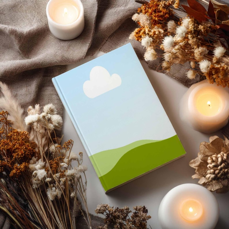 May include: A hardcover book with a sky and cloud design. The cover features a gradient blue sky, a white cloud, and green rolling hills. The book is surrounded by candles and dried flowers, creating a cozy, natural aesthetic.