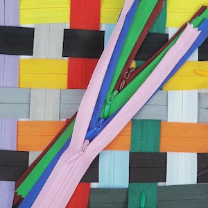 May include: A colorful assortment of zippers in various colors, including pink, blue, green, and brown, are displayed against a woven background of different colored fabric strips. The zippers are partially open, showcasing their teeth and sliders.