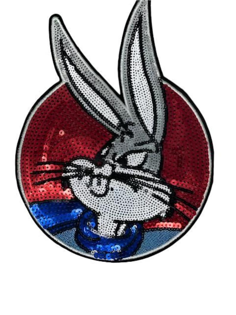 Puede incluir: Un parche de Bugs Bunny con un fondo rojo con lentejuelas. El conejo de dibujos animados es gris y blanco con detalles en negro, con un cuello azul. El parche es circular con un borde negro.