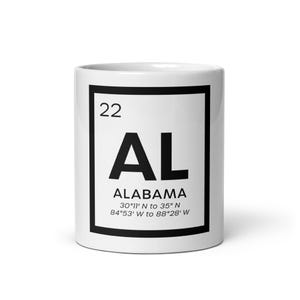 Könnte beinhalten: Eine weiße Keramiktasse mit schwarzem Rand, die den Text "ALABAMA" im Stil des Periodensystems zeigt. Die Tasse enthält auch die Koordinaten des Staates: 30°11' N bis 35° N und 84°53' W bis 88°28' W.
