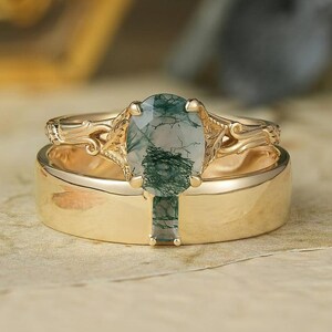 May include: A gold wedding ring set. The engagement ring features an oval-cut, green moss agate gemstone. The band has an ornate design. The wedding band is a simple, polished gold band. The rings are set on a floral patterned surface.