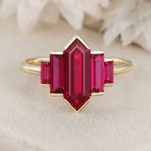 May include: A gold ring featuring a geometric design with a large, rectangular, ruby-red gemstone at the center, flanked by smaller, rectangular rubies. The ring has a polished, smooth band.