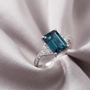 May include: A silver-toned ring featuring a large, rectangular, emerald-cut dark blue gemstone at the center. The band is accented on each side by a triangular cluster of three small, clear, round-cut stones set in a polished metal frame.