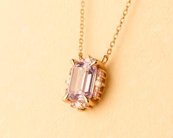 May include: A gold-toned necklace featuring a rectangular, light purple gemstone pendant. The pendant is accented with smaller pink and white stones. The chain is delicate, and the background is a soft, warm yellow.